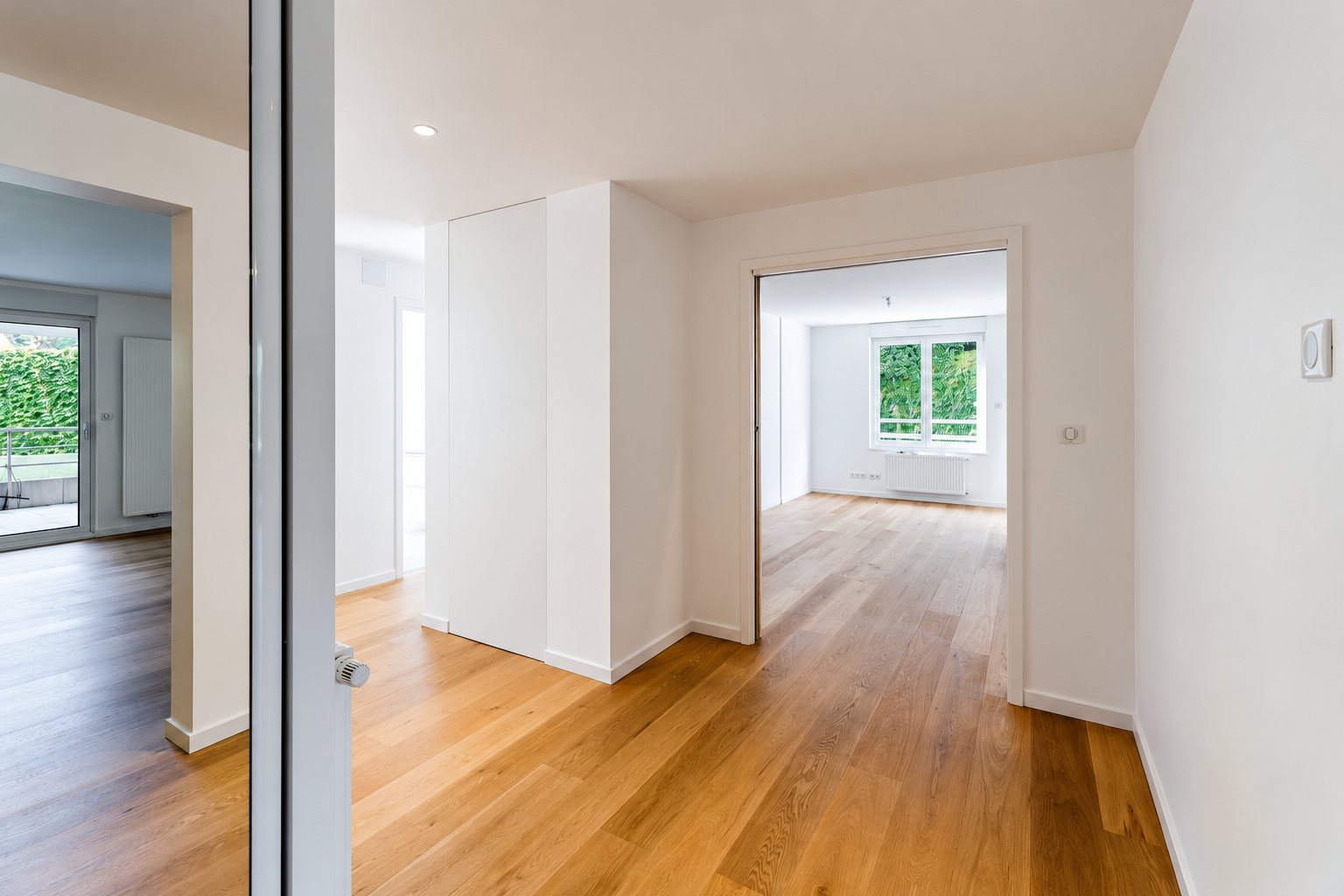 Couloir d'un appartement rénové et vide, avec parquet en chêne clair, murs blancs et vue sur un salon lumineux donnant sur la verdure.