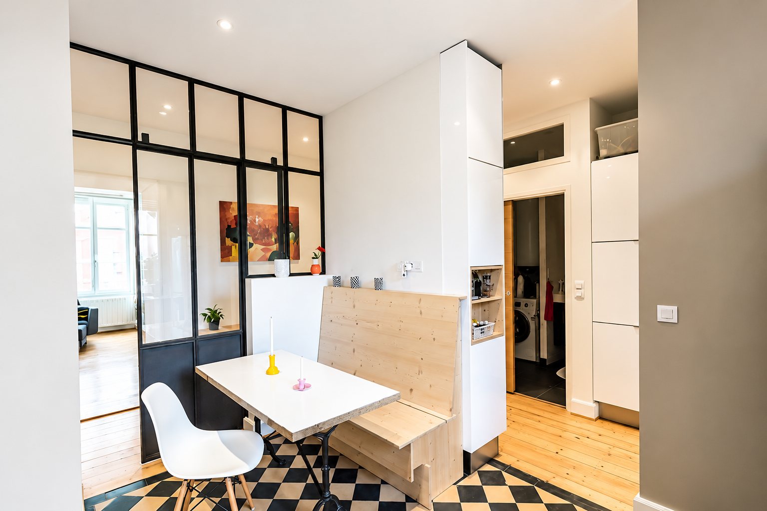Un coin repas moderne avec banquette en bois clair, table blanche, chaise design et verrière noire, sur un sol en carreaux noir et blanc.