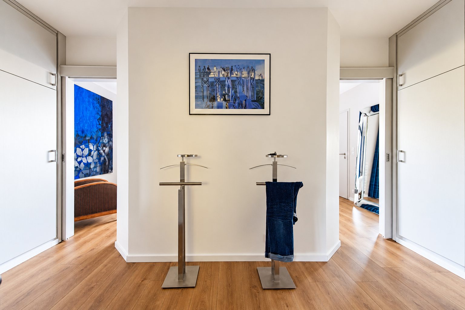 Un couloir moderne aux murs blancs avec deux valets de nuit en métal devant un tableau encadré, des placards coulissants de chaque côté et un parquet clair.