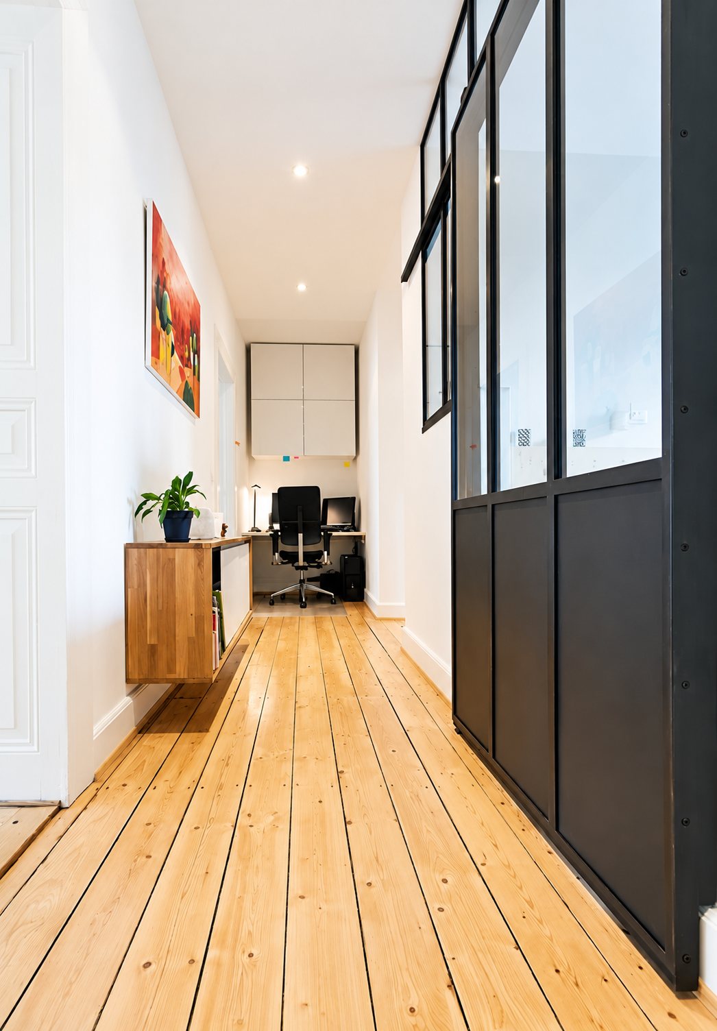 Un long couloir aux murs blancs avec un parquet clair, une verrière d'atelier noire sur la droite, un meuble en chêne suspendu et un petit bureau au fond.