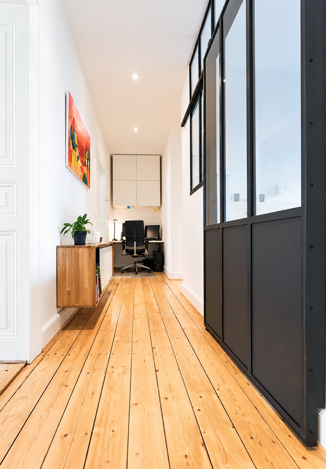 Un long couloir au parquet clair bordé d'une verrière d'atelier noire, avec un meuble en chêne suspendu et un petit bureau au fond.