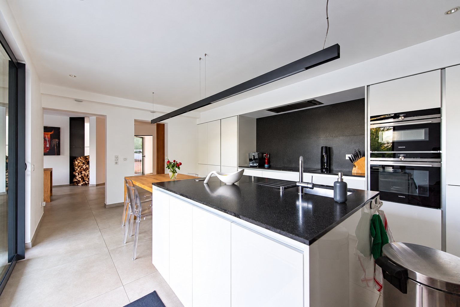 Cuisine contemporaine aux façades blanches avec un grand îlot à plan de travail noir, des fours encastrés et une salle à manger lumineuse en arrière-plan.