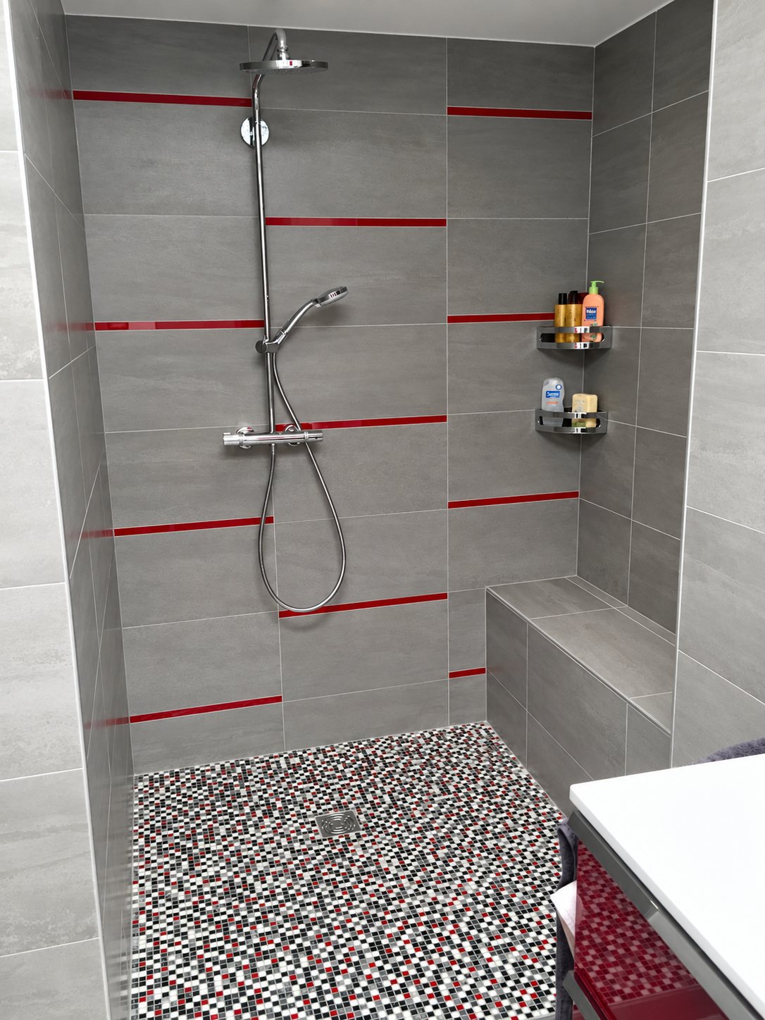 Douche à l'italienne rénovée aux murs en carrelage gris orné de fines bandes rouges, avec banc carrelé, étagères d'angle et sol en mosaïque rouge, noire et blanche.