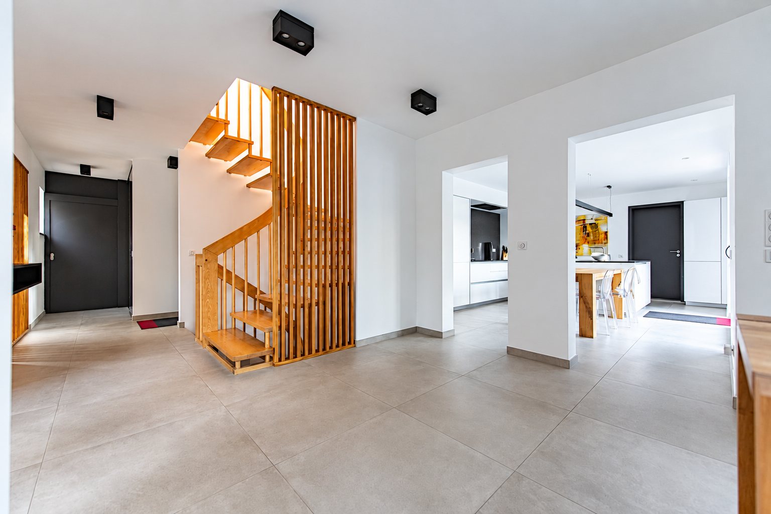 Grand hall moderne au carrelage gris clair avec un escalier en bois à claustra et, au fond, une cuisine blanche ouverte.