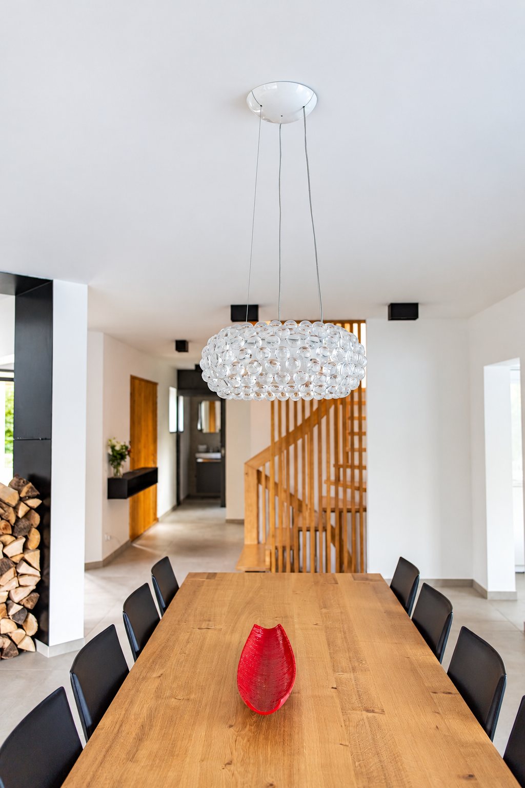 Une grande table en bois clair entourée de chaises noires, ornée d'un vase rouge et surmontée d'un lustre en bulles de verre, dans un intérieur lumineux avec escalier en bois.