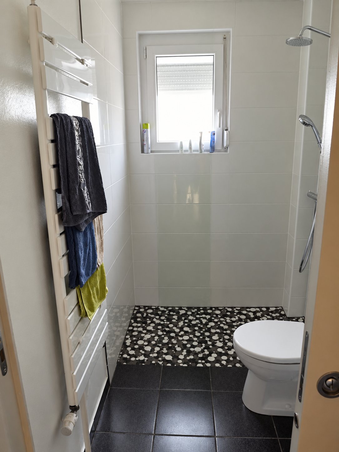 Une salle de bains étroite aux murs carrelés de blanc avec une douche à l'italienne au sol de galets noirs et blancs, un sèche-serviettes et des toilettes.
