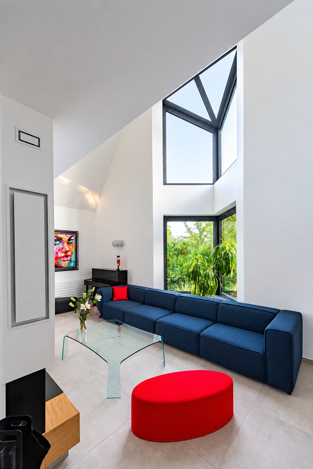 Salon contemporain à double hauteur avec un grand canapé bleu, un pouf rouge, une table basse en verre et de grandes fenêtres donnant sur la verdure.