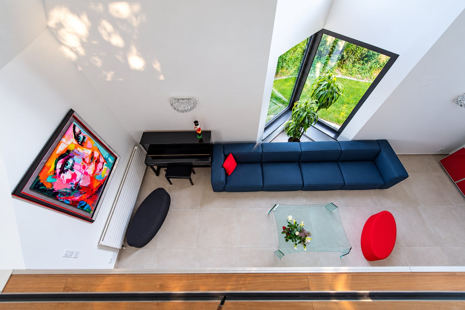 Vue plongeante sur un salon lumineux avec un grand canapé bleu, une table basse en verre, un piano noir et un tableau coloré au mur.