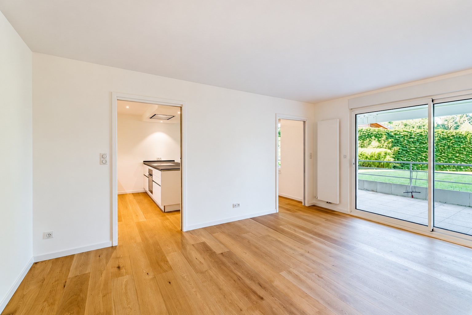 Pièce de vie vide aux murs blancs et au parquet clair, avec une cuisine moderne en arrière-plan et une grande baie vitrée donnant sur une terrasse et un jardin.